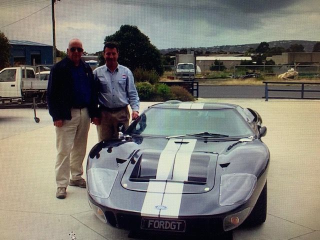Two men standing beside a silver-grey GT40 with white centre stripes in an industrial yard